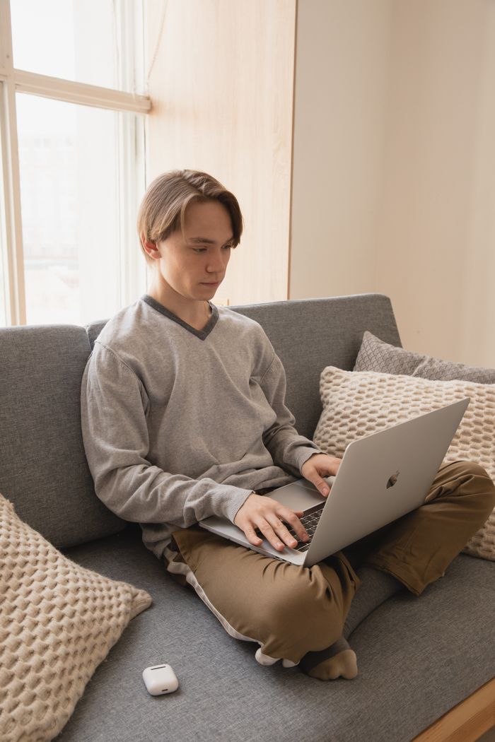 contact-img Young male remote worker siting with crossed legs on cozy sofa with cushions near window while typing on netbook in flat